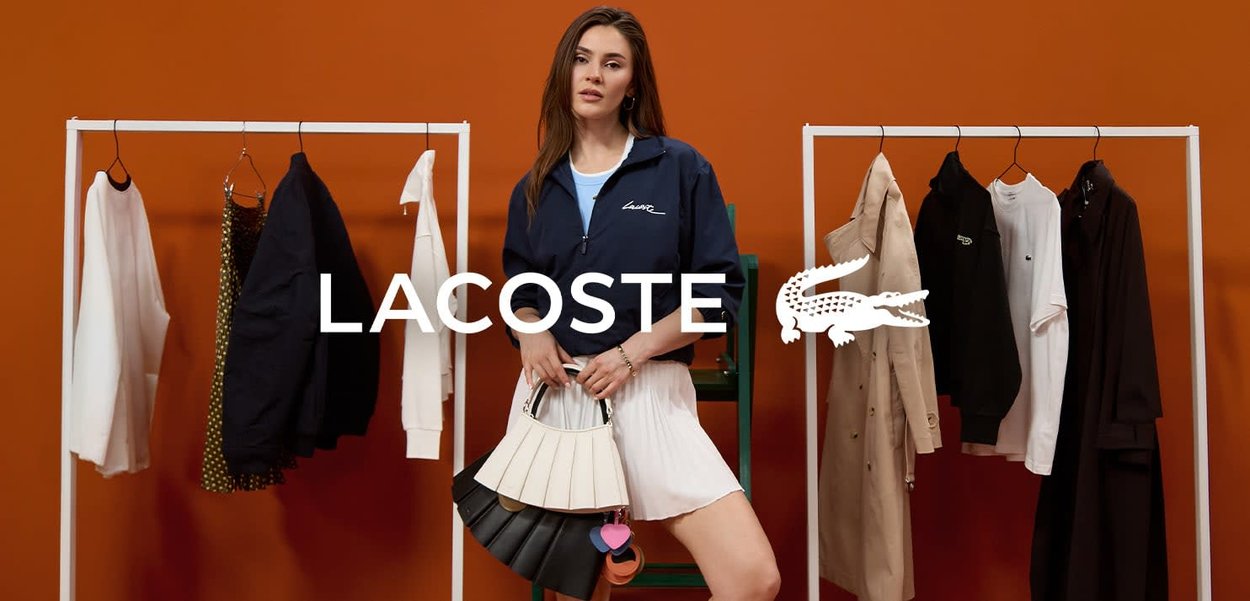 Mujer con chaqueta deportiva y falda blanca sostiene bolsos, situada entre dos percheros contra un fondo naranja con el texto y logotipo de "Lacoste".