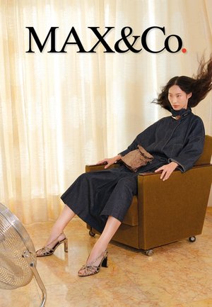 Femme aux longs cheveux rejetés en arrière, assise dans un fauteuil marron, portant une tenue en denim foncé et des chaussures à talons hauts imprimées, près d'un ventilateur argenté.