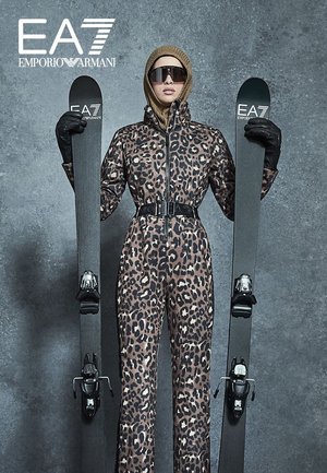 Combinaison de ski imprimé léopard avec une fermeture éclair à l'avant et une ceinture, portée avec des gants noirs. Le modèle tient des skis noirs avec des fixations de marque sur un fond gris.