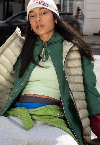 Groene pufferjack over een mintgroene top. Grijze sweatpants met een geribbelde groene riem. Beige beanie met rode accentletters.