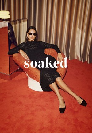 Schwarzes geripptes Kleid, orangefarbener Plüschstuhl, glatte weiße Basis, schmaler Absatz schwarzer Schuhe. Reflektierte Figur auf einer glänzenden Oberfläche, warmfarbiger Hintergrund.