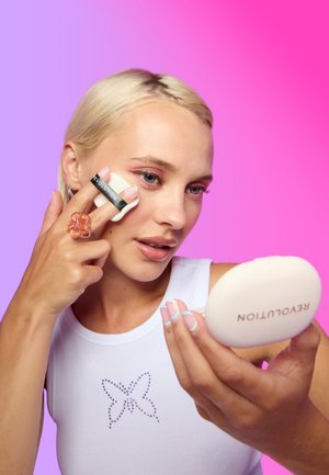 Blonde Frau trägt mit einem Schwamm Make-up auf ihre Wange auf, während sie einen Kompaktspiegel mit der Aufschrift "Revolution" vor einem rosa-lila Hintergrund hält.
