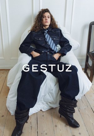 Vrouw met krullend haar die achterover leunt op een witte zitzak, gekleed in een donker denim outfit, gestreepte stropdas en losse zwarte laarzen op een houten vloer.