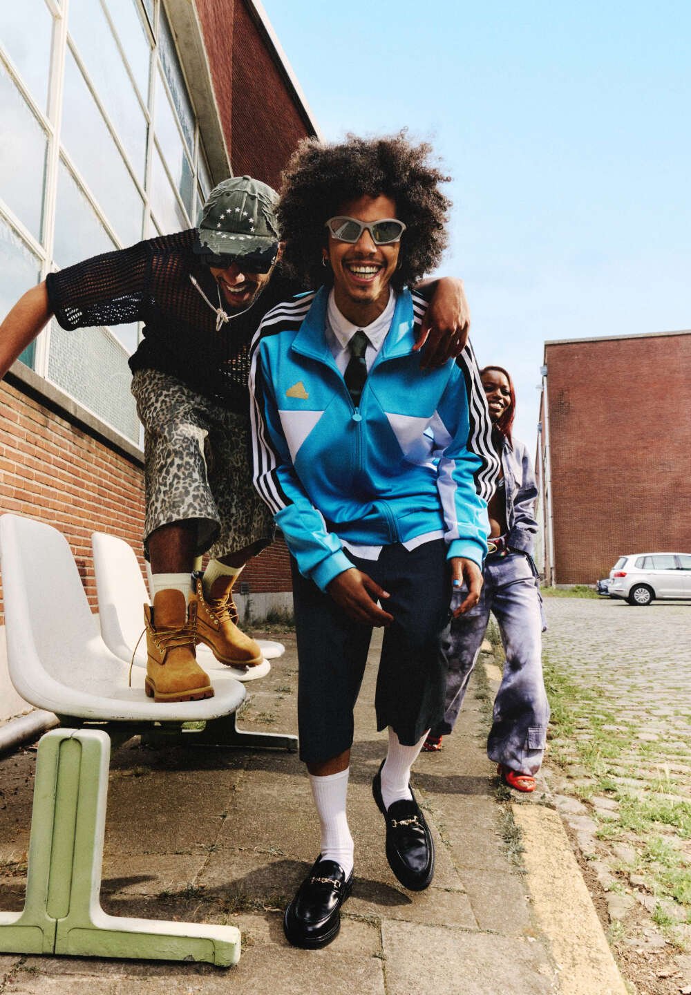 Blauwe trainingsuit met witte accenten, zwarte loafers, lichtgrijze shorts en tanlaarzen. Drie mensen poseren buiten nabij witte stoelen en kasseien.