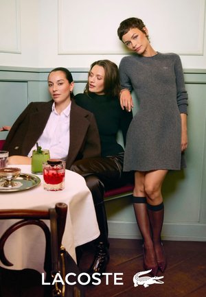 Vêtements Lacoste sur trois femmes : l'une en blazer marron sur une chemise blanche, une autre en haut noir, et la dernière en robe grise, avec des boissons sur la table.
