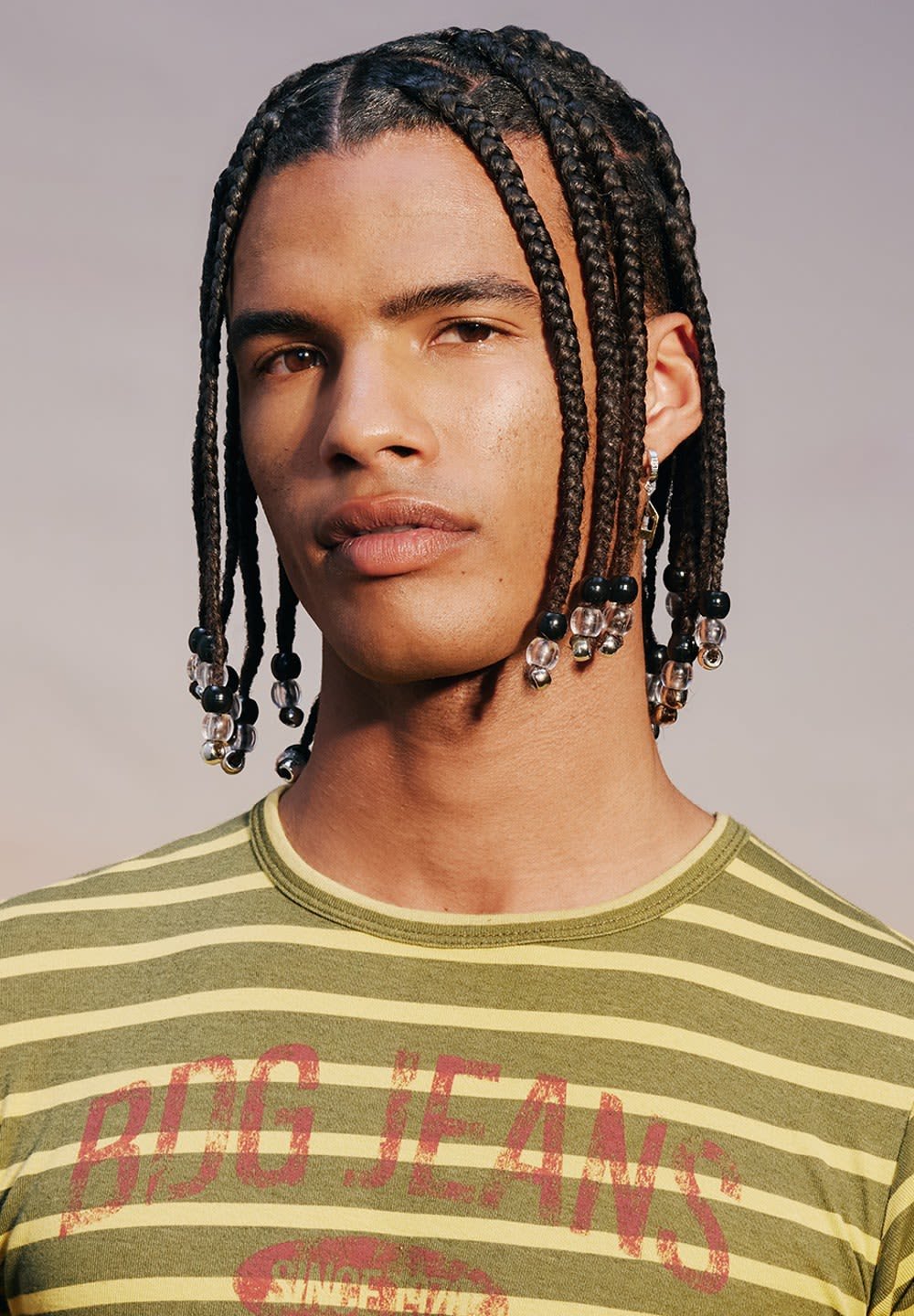 Modèle portant un t-shirt rayé vert avec "BDG JEANS" imprimé en rouge. Cheveux coiffés en longues tresses avec des accents de perles. Texture de peau naturelle visible.