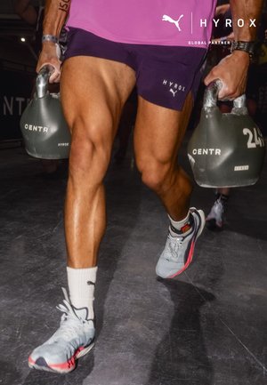 Athlet trägt lila Shorts und graue Sneakers und hält während einer HYROX-Fitnessveranstaltung zwei Kettlebells mit der Aufschrift CENTR.