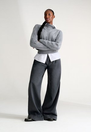 Maglione grigio a collo alto in maglia con texture a coste, indossato sopra una camicia bianca a bottoni. Pantaloni larghi grigio scuro a gamba larga e morbidamente aderenti.