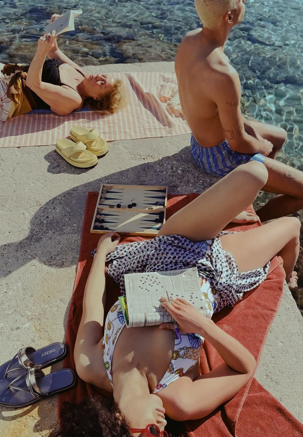 Grupa ljudi opušta se na kamenitoj površini blizu vode. Ovdje su vidljive šarene kupaće kostime, sandale i set za backgammon.