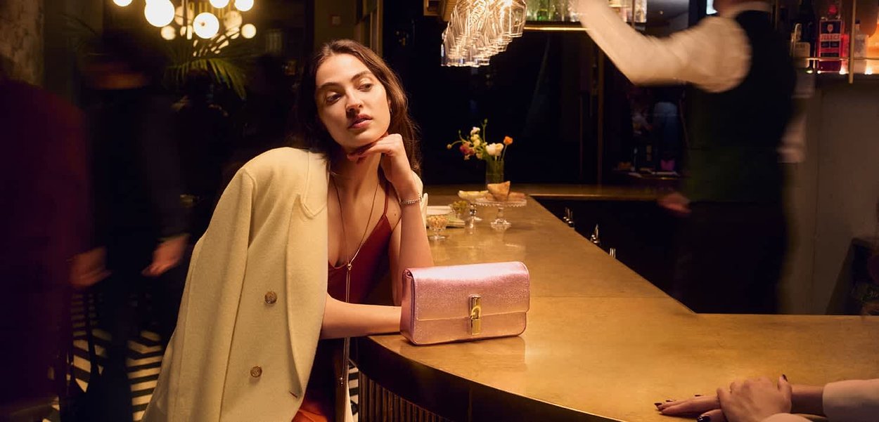 Metallic pink clutch bag with a clasp closure, positioned on a bar countertop; smooth texture, rectangular shape, and prominent sheen.