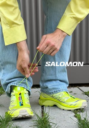 Person i blå jeans og gul skjorte binder lysegule Salomon-sneakers udendørs på et brolagt område med græs. Salomon-logoet er synligt.