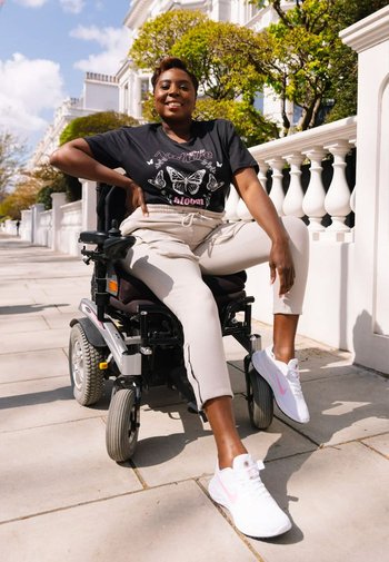 Svart grafisk t-shirt med fjärilstryck, beige sweatpants med sidosträck, vita sneakers med rosa accenter, och en elektrisk rullstol.