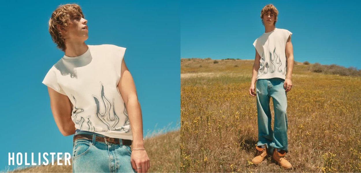 Jonge man in een mouwloos wit shirt met vlammenpatroon en blauwe jeans staat in een droog grasveld onder een heldere blauwe lucht.