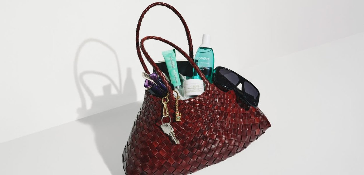 Dunkelrote gewebte Handtasche mit Sonnenbrille, Schlüsseln und verschiedenen Kosmetika auf weißem Hintergrund.