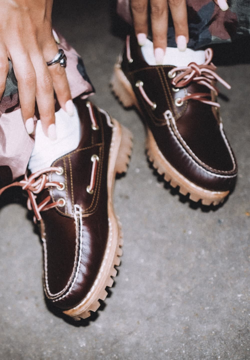 Hände mit rosa Nägeln passen dunkelbraune Lederschuhe mit dickem beigen Absatz und hellrosa Schnürsenkeln auf einem Betonboden an.