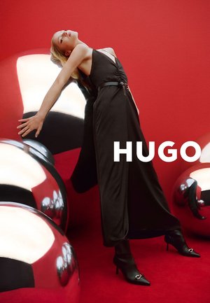 Frau im schwarzen Kleid und mit Absätzen lehnt sich zurück an eine große reflektierende silberne Kugel auf rotem Boden und Hintergrund mit dem Schriftzug „HUGO“.