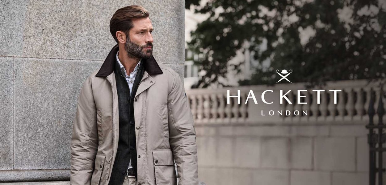 Hombre con barba, vestido con chaqueta beige sobre blazer oscuro y camisa a cuadros, de pie junto a una pared de piedra al aire libre cerca del logo de Hackett London.