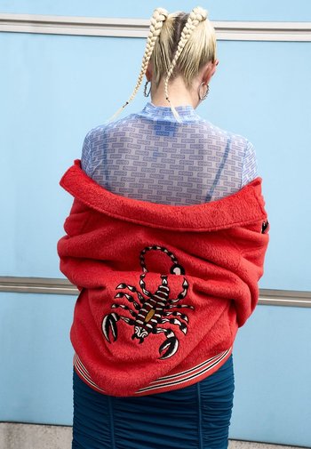 Rode fleecejas met een schorpioenborduursel op de rug. Draagt over een blauw top met patroon. De outfit bestaat uit een geribbelde, aansluitende rok.