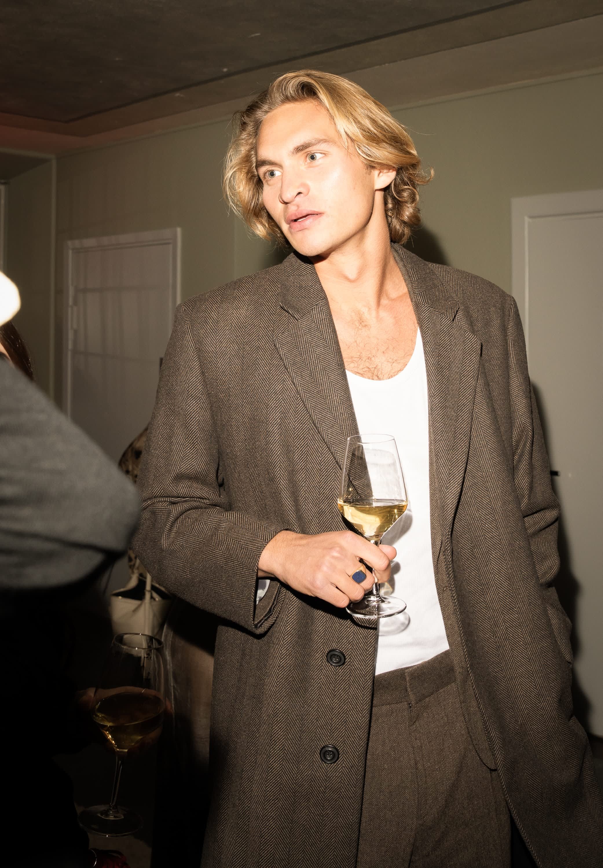 Brun fiskbensmönstrad överrock bärs över en vit skjorta, håller ett klart vinglas med en ljus dryck. Kort, vågigt blont hår.