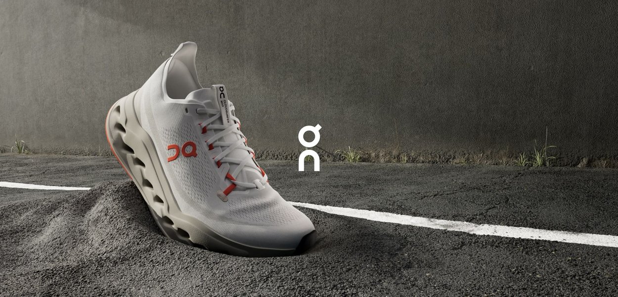 Weiße Sportschuhe mit einem Obermaterial aus Mesh, runder Form, auffälligen seitlichen Ausschnitten und Markenlogo. Auf einem strukturierten dunklen Hintergrund platziert.