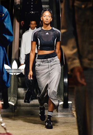 Woman walks down runway wearing black Adidas crop top, gray skirt, black sneakers, and carrying a small black handbag, with people behind.