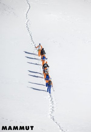 Sieben Wanderer in orange und blauer Ausrüstung wandern in Reihe durch tiefen Schnee und hinterlassen eine Spur über eine weite, weiße Landschaft.