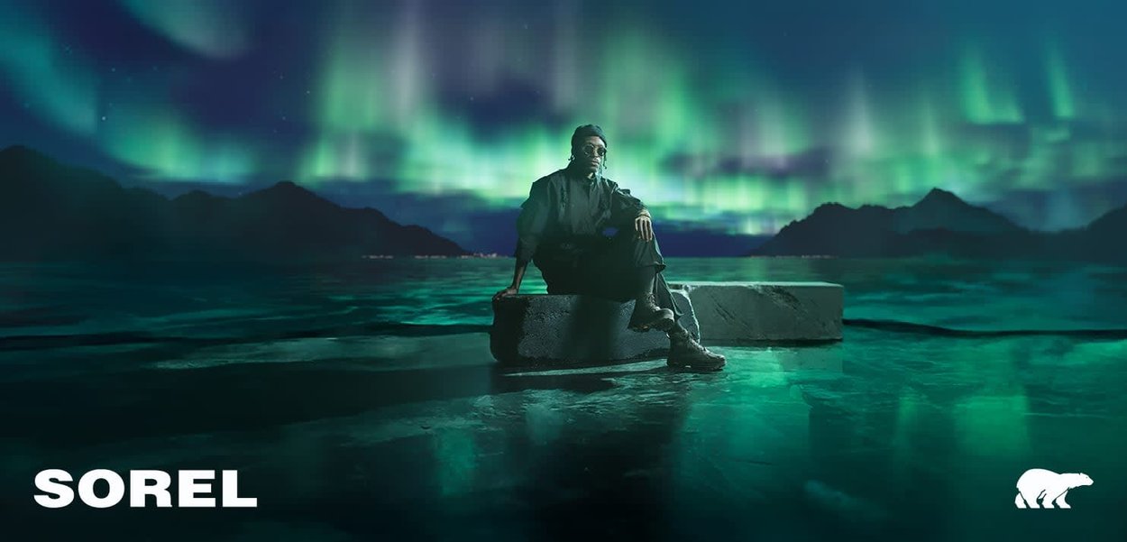 Sorel Markenfußbekleidung vor einem Aurora-Hintergrund präsentiert. Dunkles Obermaterial, robustes Design mit einer klobigen Sohle. Coole Farbtöne.