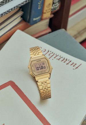 Reloj de pulsera digital Casio dorado sobre un libro con texto visible y una pila de libros en el fondo.