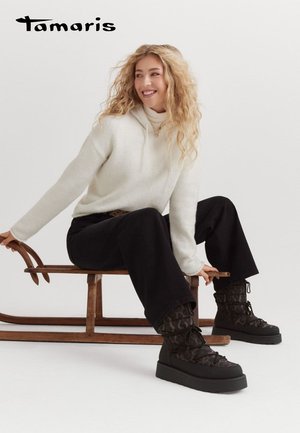 Haut à col montant blanc associé à un pantalon noir à larges jambes et des bottes à lacets noires à motifs sur un traîneau en bois.