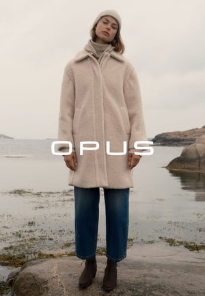 Lichtbeige, fluffy jas met een kraag en voorzakken, gecombineerd met blauwe wijde jeans en bruine enkellaarzen. Het model draagt een gebreide muts.