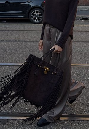 Personne portant un pantalon large marron et un pullover sombre, tenant un grand sac fourre-tout noir à franges tout en marchant dans la rue.