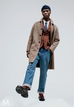 Manteau en pied-de-cochon marron, superposé à un blazer marron et une chemise blanche, jean bleu, bottes à lacets noires avec des accents rouges, bonnet bleu marine.