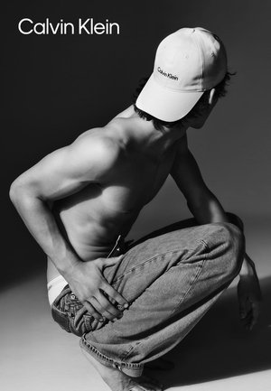 Weißer Baseballcap mit "Calvin Klein"-Logo, nackter Oberkörper, Jeans, hockende Pose, einfarbiger Hintergrund, sanfte Schatten.