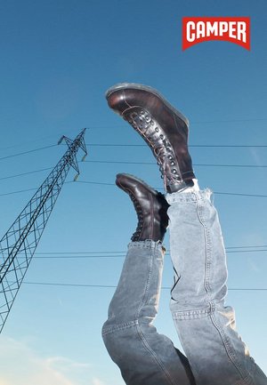 Legs in light gray jeans and dark brown lace-up boots raised against a blue sky with a power line tower and Camper brand logo.