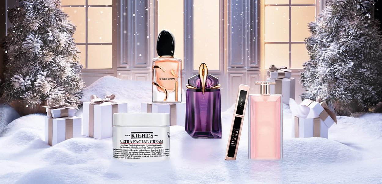 Four perfume bottles in varying shapes and colors, plus a Kiehl's facial cream; snowy background with wrapped gifts and a Christmas tree.