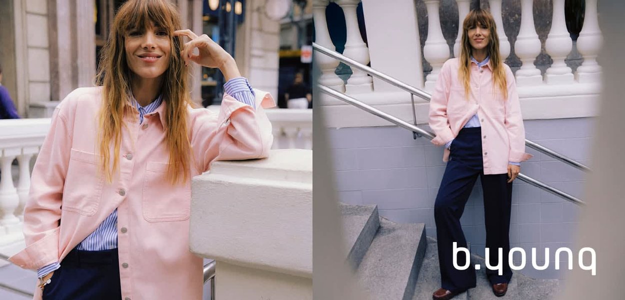 Hellrosa übergroße Jacke, blau gestreiftes Hemd darunter, dunkelblaue Hose und braune Schuhe, posiert nahe einem weißen Geländer und Treppen.