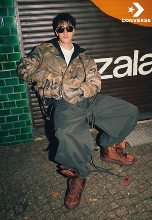 Chaqueta bomber de camuflaje, pantalones oscuros oversize, botas altas marrones y gafas de sol. Situado frente a una pared verde con azulejos y un fondo de grafiti.