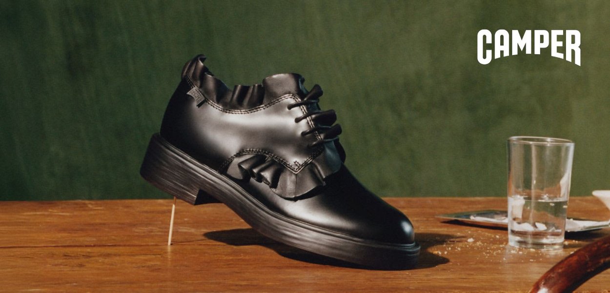 Czarny skórzany but na sznurowadła z ozdobnym marszczeniem, spoczywający na drewnianej powierzchni, obok szklanka wody i podkładka, logo "CAMPER" w prawym górnym rogu.