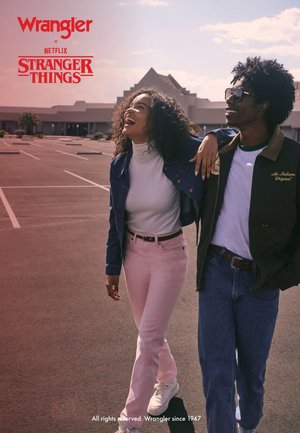 Paar in lässiger Kleidung: Frau in weißem Rollkragenpullover, Jeansjacke und pinken Jeans; Mann in Jacke mit grünem Besatz und Sonnenbrille. Im Hintergrund ein leerer Parkplatz.