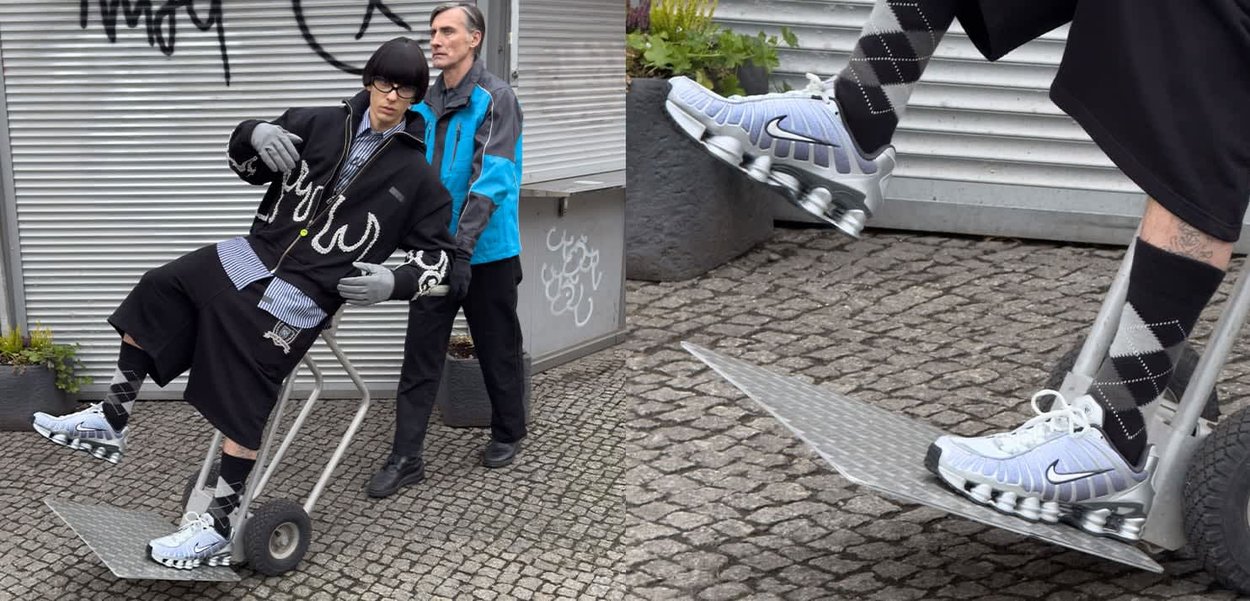 Nike-sko i lys grå med sølvfarvede accenter, båret sammen med sorte shorts og mønstrede sokker, er vist på en vogn med hjul foran en tekstureret overflade.