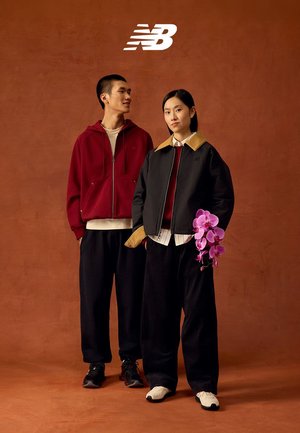 Zwei junge Erwachsene in lässiger New Balance Kleidung stehen nebeneinander vor einem braunen Hintergrund; einer hält eine pinkfarbene Orchidee.