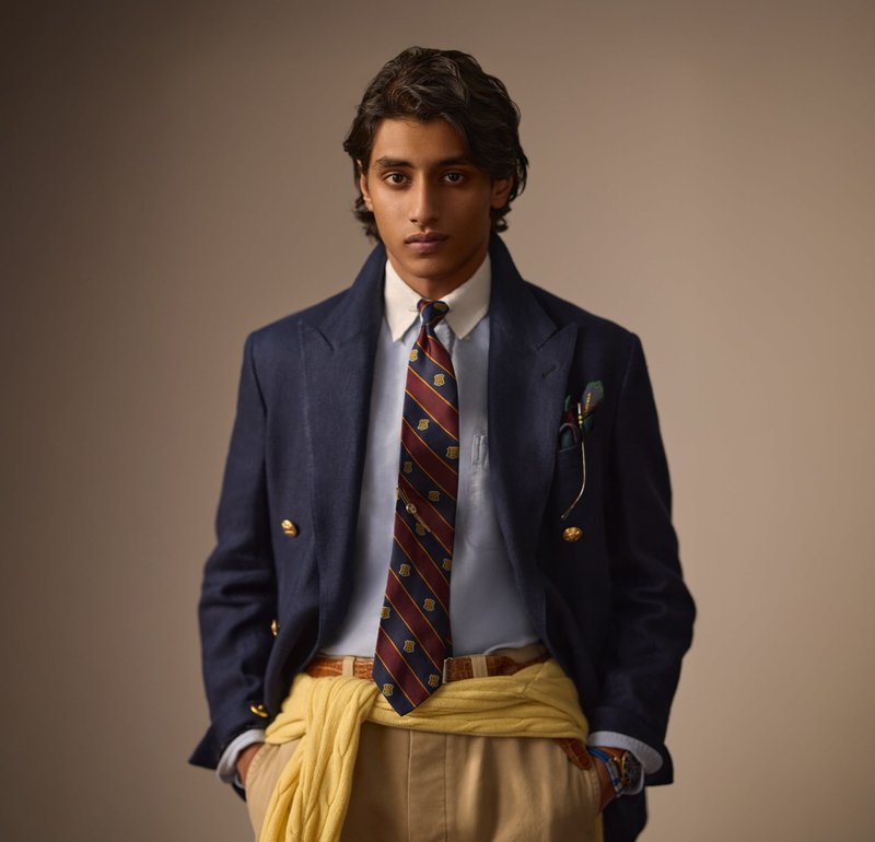 Marineblå dobbeltbrystet blazer, hvid skjorte med krave, stribet slips med guld accenter, gul sweater bundet om livet, beige bukser, brun bælte.