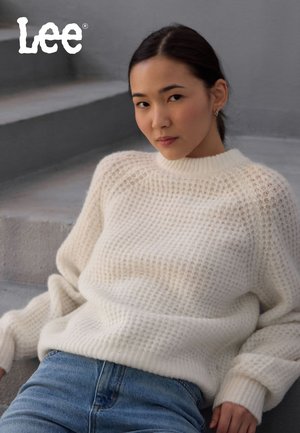 Kremfarget strikkegenser med løs passform, som har et strukturert mønster og ribbestrikket krage. Sammen med blå jeans.