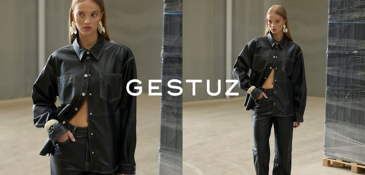 Kvinde i oversize sort læderskjorte og bukser, poserende i minimalistisk lagerområde med indpakkede paller, mærket "GESTUZ" centreret.