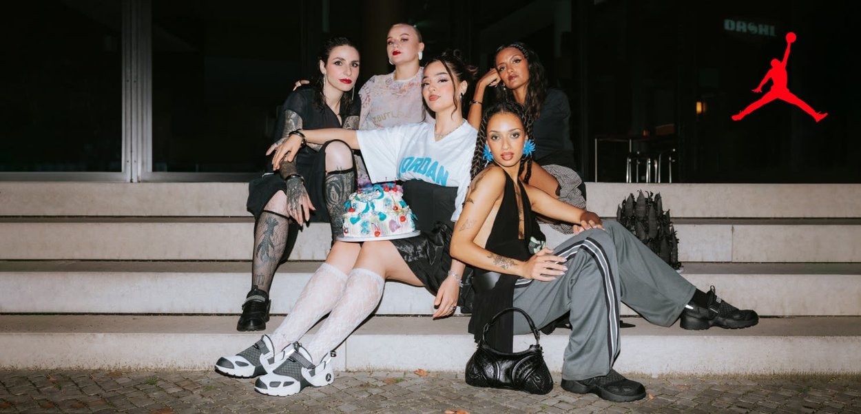 Five individuals sit on stairs. Varied outfits include a white jersey, fishnet tights, and sleek handbags. A cake with colorful accents is present.