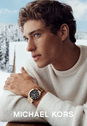 Goldfarbene Metallarmbanduhr mit schwarzem Zifferblatt, römischen Ziffern und Gliederarmband. Das Modell trägt einen strukturierten weißen Pullover vor einem verschneiten Hintergrund.