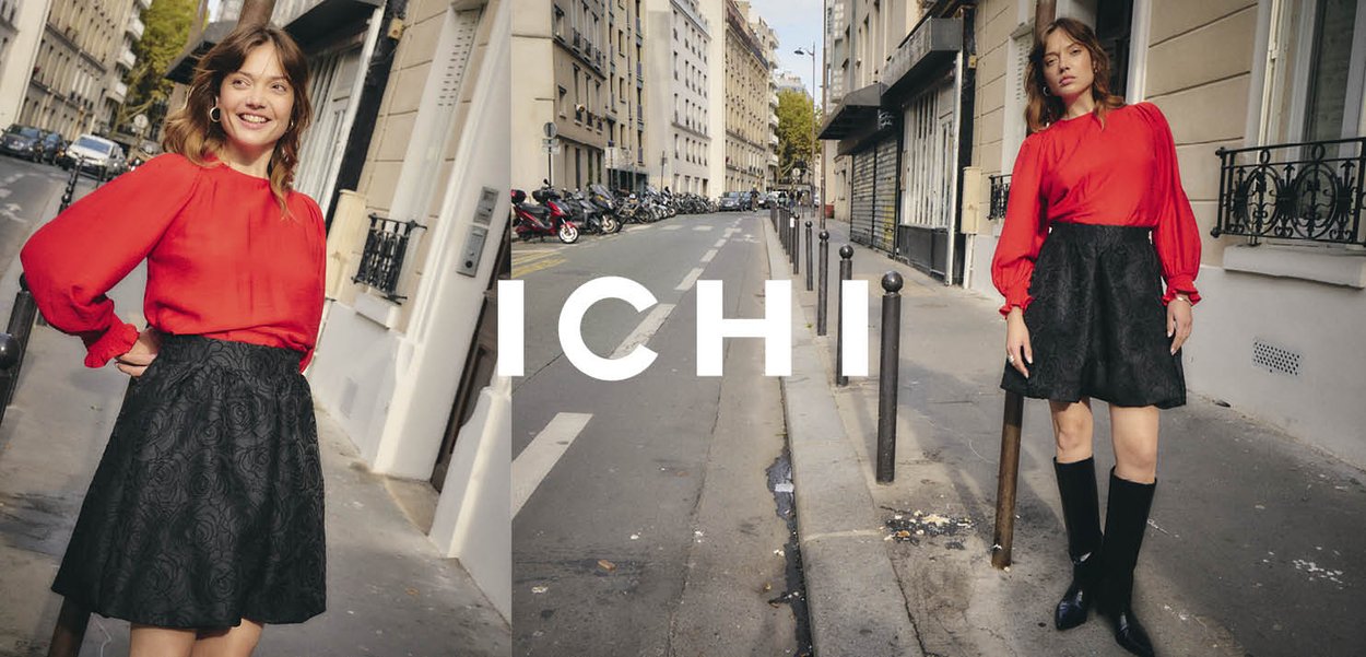 Rote Bluse mit langen Ärmeln und gerafften Schultern, kombiniert mit einem schwarzen Rock mit floralem Muster, ergänzt durch kniehohe schwarze Stiefel.