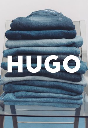 Pila di jeans in denim in diverse tonalità di blu, dal blu scuro al lavaggio chiaro, piegati ordinatamente su un tavolo espositivo in vetro. Logo del marchio visibile.