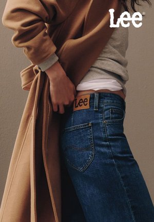 Donkere blauwe spijkerbroek met een bruin label waarop "Lee" staat. Gecombineerd met een grijze trui en een lange beige jas, wat een casual gelaagde look laat zien.