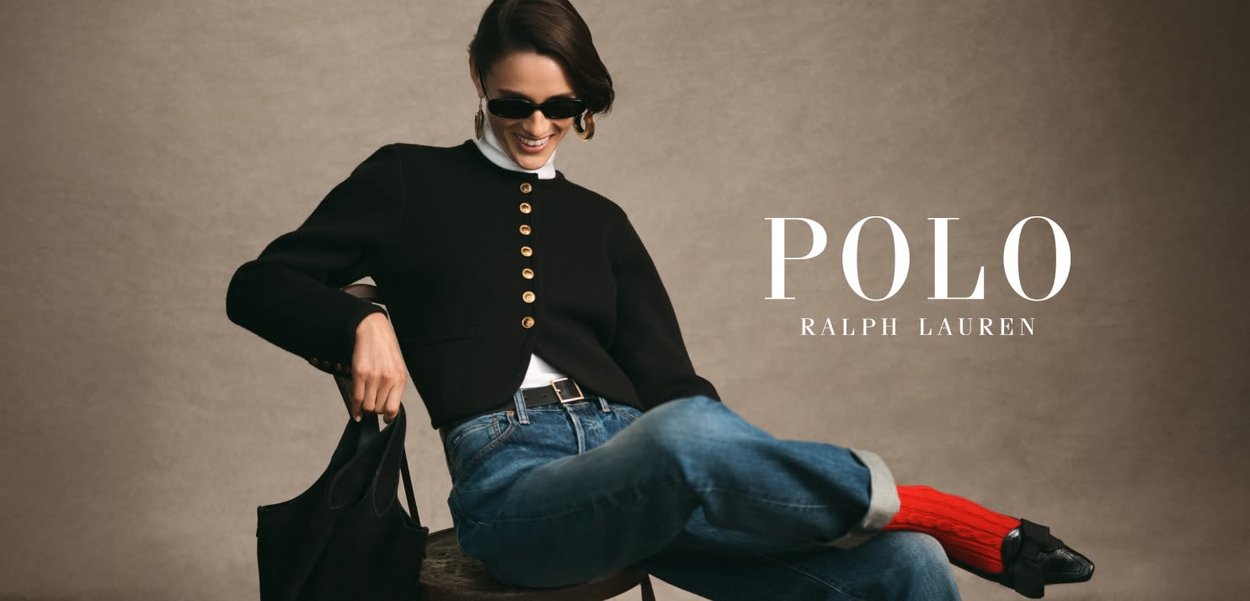 Black button-up cardigan, white turtleneck, blue jeans, red socks, black shoes, holding a black bag, seated on a wooden stool with a gray background.